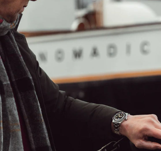 Person in coat and scarf next to a boat named NOMADIC, showing their wristwatch.