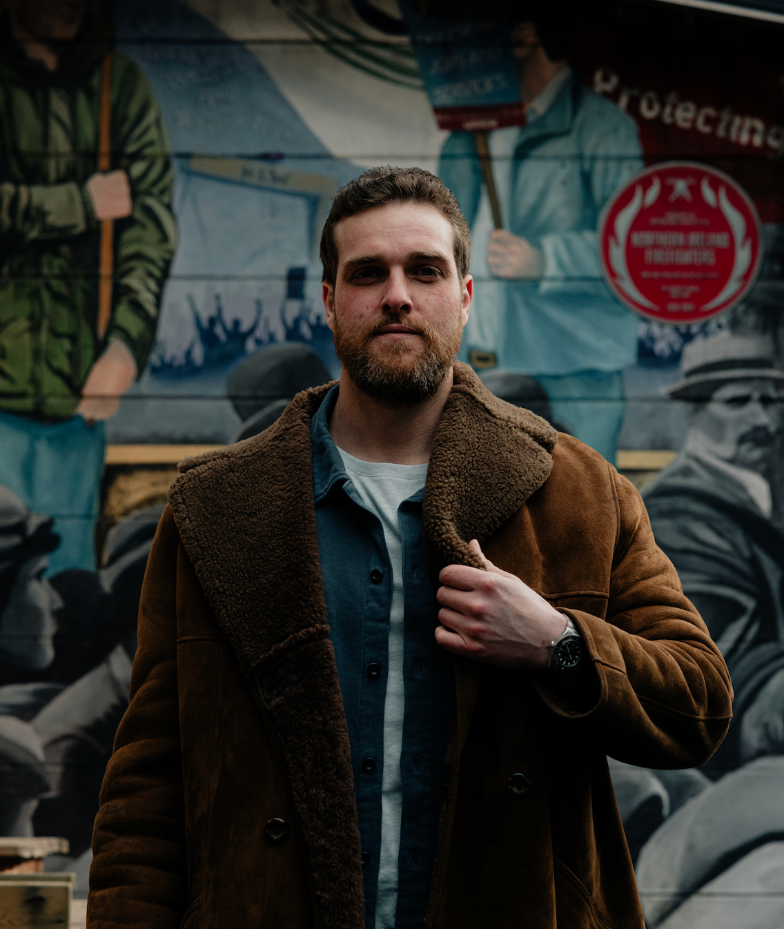 A person standing in front of a colorful mural, wearing a brown coat.